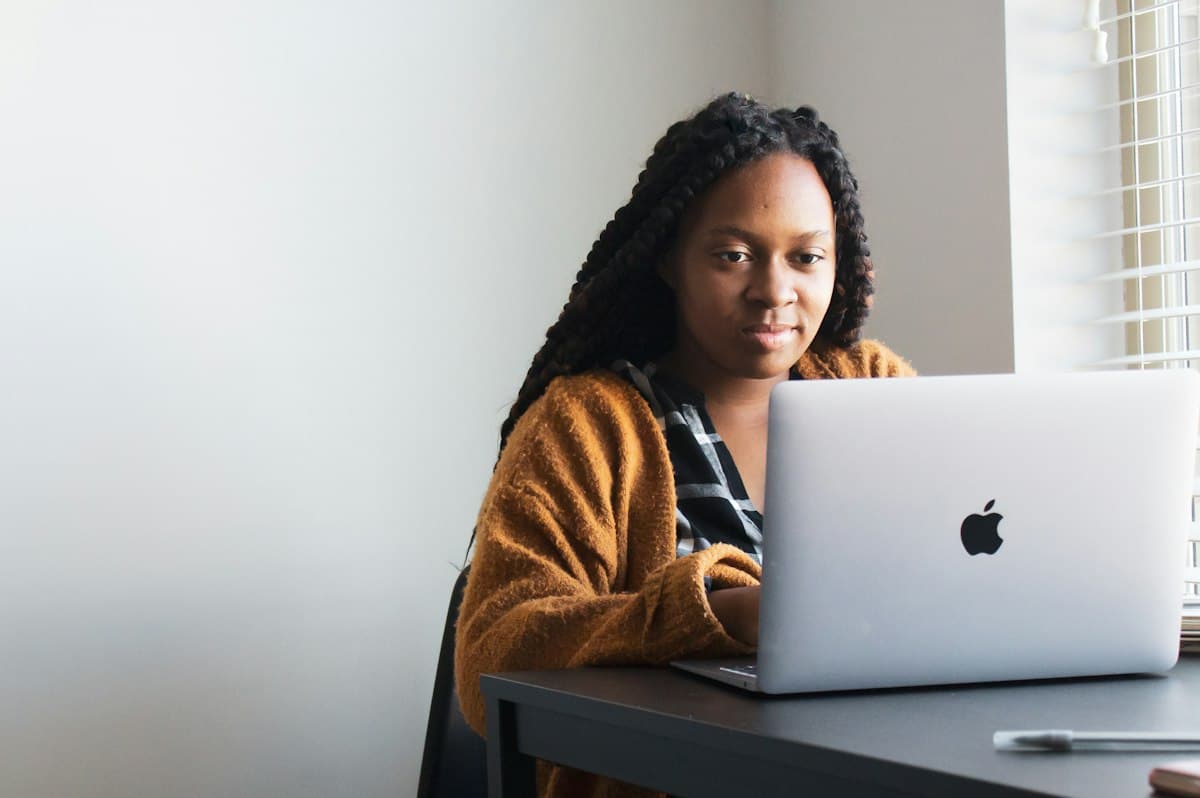 A woman with long braids works on a laptop at a desk by a bright window